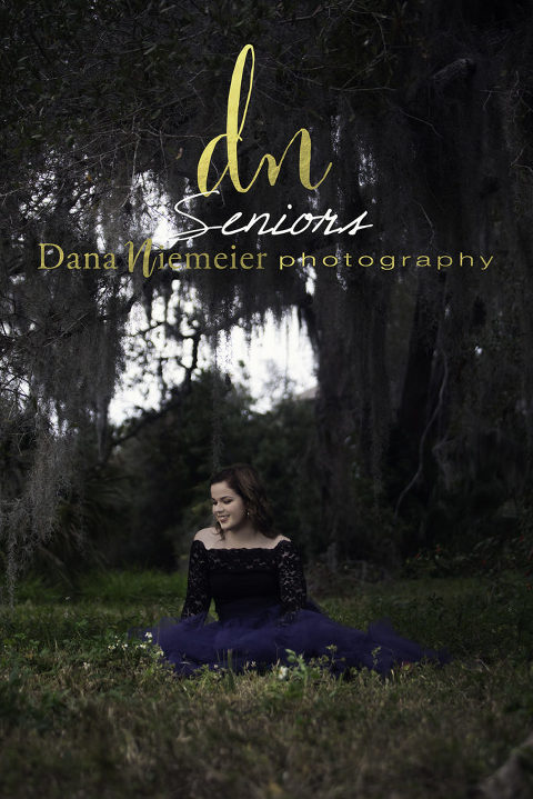 Smiling 18 year old brown hair young lady in black lace off shoulder top and a royal blue long tulle skirt sitting on the ground under an oak tree with hanging Spanish moss