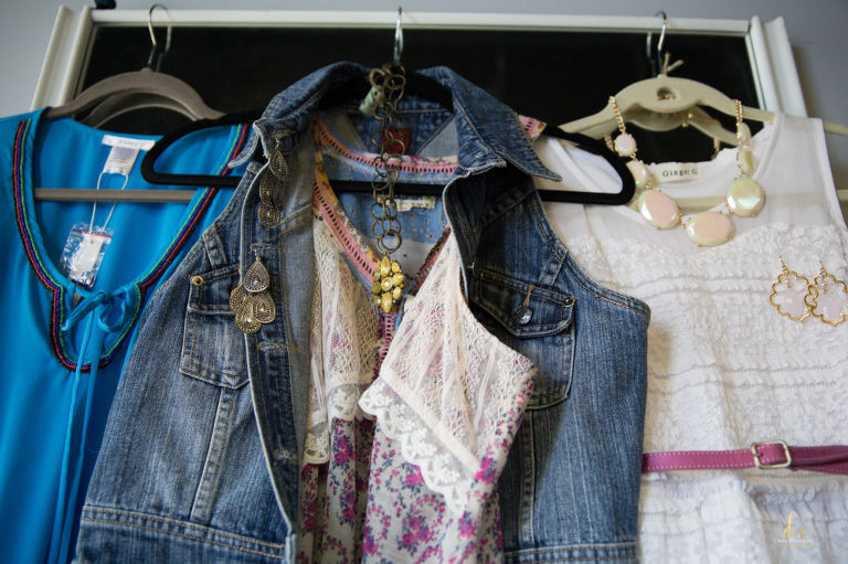 3 clothes hangers with outfits, blue dress, jean vest and yellow necklace and white lace dress with thin pink belt and opalescent large stone necklace