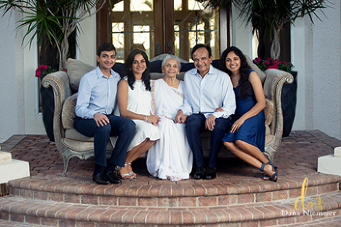 Family sitting on beige velvet sofa on raised brick tiled platform in front of a double door with arched entry. Small palms arching over doorway with light streaming through the glassed doors South East Asian family in blues, white and cream, smiling sitting close together