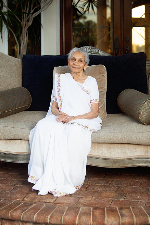 Older grandmother sitting on beige velvet couch wearing a white sari hands clasped in her lap.